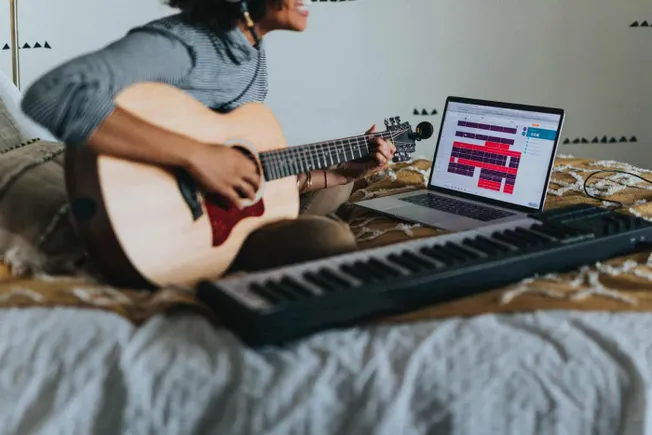 Guitarist creating music using a laptop, keyboard, and acoustic guitar in a home studio setup for digital music production an