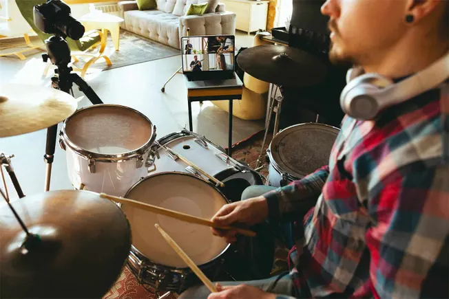 Professional drummer participating in virtual band rehearsal on laptop, creative home studio setup with drums, headphones, an