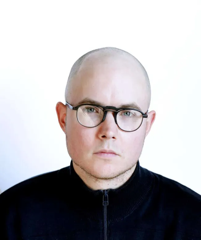 Bald man wearing black glasses and black zip-up jacket against white background, close-up portrait.