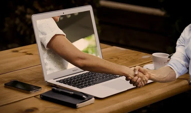 Handshake between a person on a laptop screen and another person in real life, symbolizing virtual collaboration or online pa