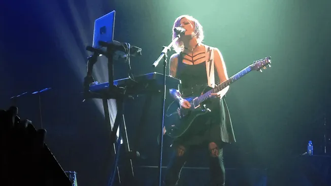 Young female musician performing live with electric guitar and keyboard on stage under dramatic lighting.