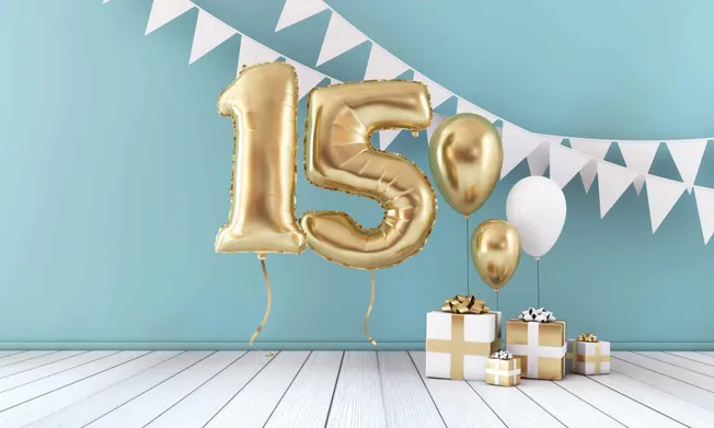 15th anniversary celebration with gold number balloons, white and gold gift boxes, and festive banners on a light blue wall b