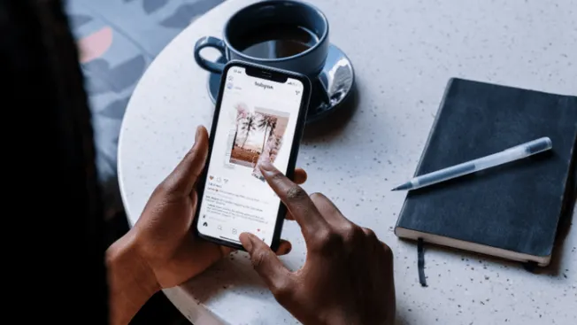 Mobile phone displaying Instagram post with a coffee cup, notebook, and pen on a table, illustrating social media engagement