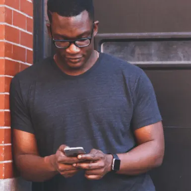 Young man using smartphone outdoors, wearing casual clothing and glasses, urban background.