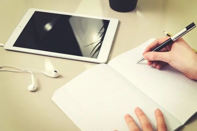 Digital tablet with blank screen and earphones on workspace, writing in graph paper notebook, productivity, tech workspace, o