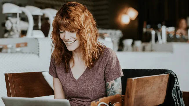 Young woman working on laptop at a cafe, smiling, with music equipment and cozy atmosphere in the background.