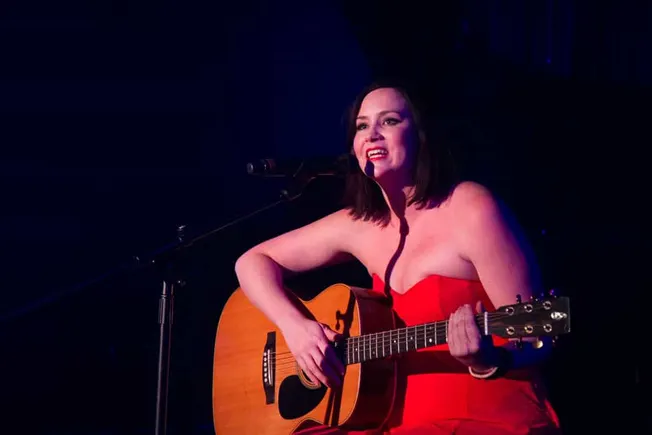 A female musician performing live on stage with an acoustic guitar, singing into a microphone, wearing a striking red dress,