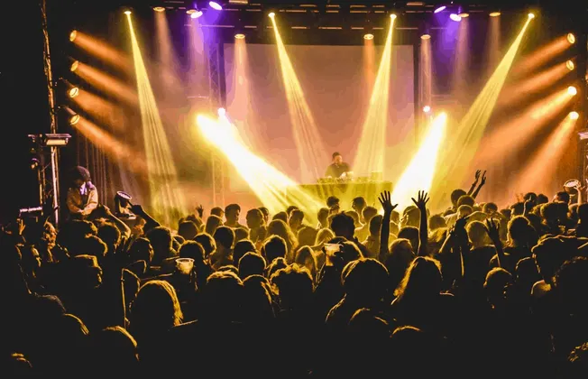 Live concert with DJ performing on stage, vibrant colorful lighting, and an enthusiastic crowd enjoying the music.