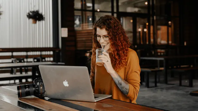 Stylish woman with curly red hair working on a laptop in a modern coffee shop, drinking iced coffee, professional digital con