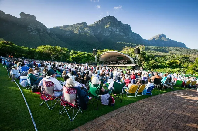 People attending an outdoor concert or event with a mountain backdrop, seated on folding chairs and enjoying live music under
