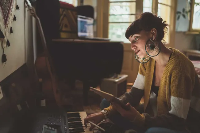 Creative musician woman playing digital keyboard and reading tablet in cozy home studio with natural sunlight.
