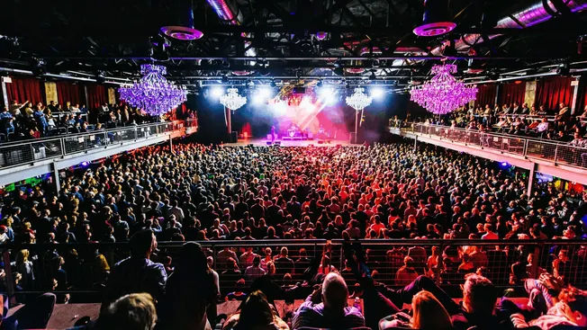 Vibrant concert hall filled with a large crowd, colorful stage lights, and elegant chandeliers, showcasing live music perform