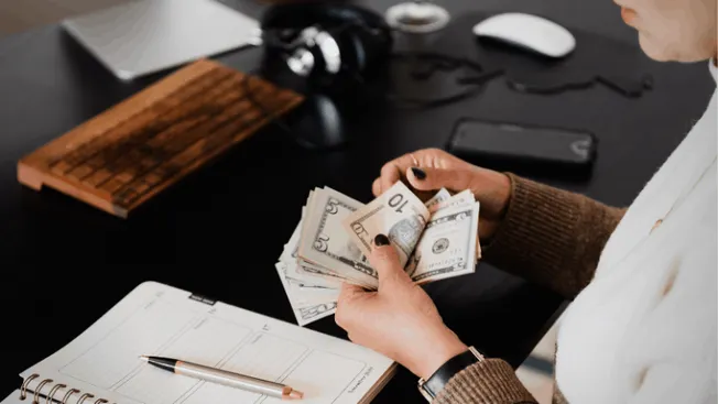 $50, $20, and $10 bills held in person's hands on desk with notebook, pen, headphones, and electronic devices, representing m