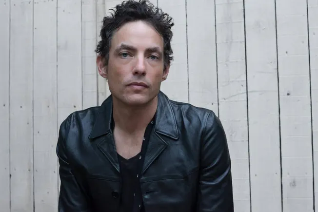 Male musician in a black leather jacket against a white wooden wall, promoting music industry insights and updates.