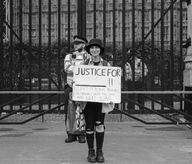 Protestor holding a "Justice For" sign at a demonstration, in front of a metal fence, highlighting social activism and public