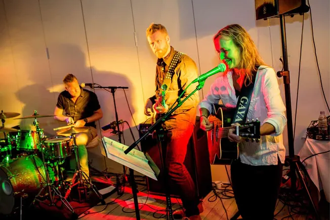 Hypebot, live musical performance featuring a guitarist and female singer with a band drummer in the background, vibrant stag