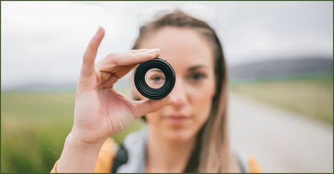 A woman holding a camera lens towards the camera, outdoor landscape background, symbolizing photography, creativity, and visu