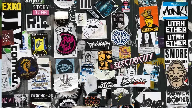Sticker collage on urban wall, featuring music, entertainment, and street art stickers, promoting various bands, brands, and