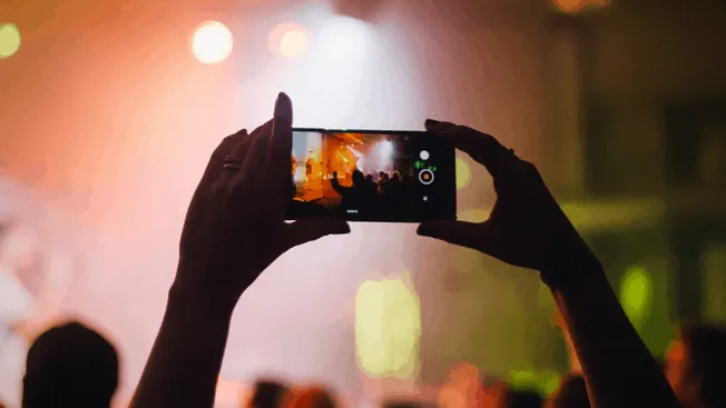 A person capturing a live concert or music event on their smartphone, with the stage and vibrant lighting visible on the scre