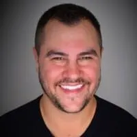 Smiling man with dark hair and beard, wearing a black shirt against a gray background, representing professionalism and appro