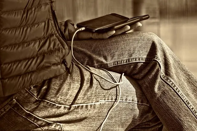 Phone charging in someone's hand while sitting on a couch with casual jeans and a padded jacket, focusing on technology and l