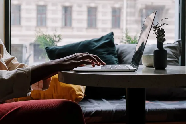 Laptop user working remotely at a cozy cafe during daytime, with a cityscape window view and minimalistic décor.