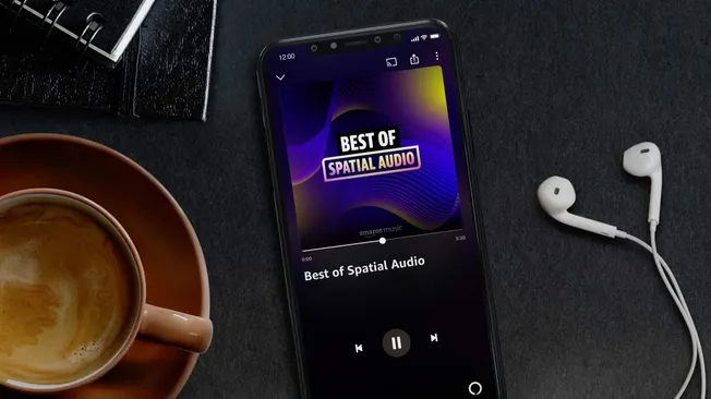A smartphone playing "Best of Spatial Audio" playlist with earbuds, coffee mug, and notebook on a dark surface, highlighting