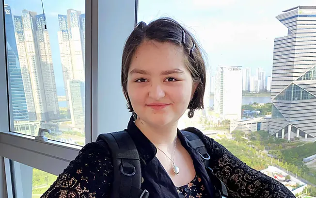 Young woman with short dark hair and earrings smiling in front of a cityscape with modern skyscrapers during daytime.