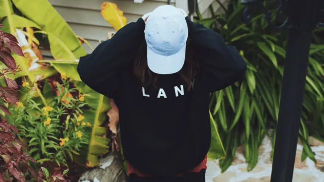 Girl wearing black sweatshirt and light blue cap among lush greenery, outdoors, lifestyle shot for fashion and casual wear ar