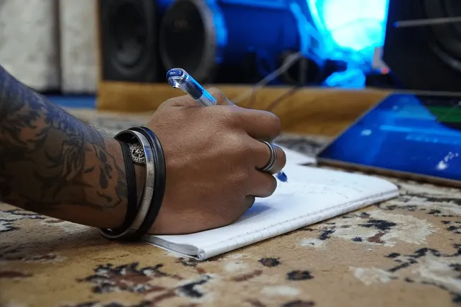 Hand writing in a notebook at a music studio or production workspace with audio equipment and speakers, representing music pr
