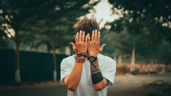 Young man covering face with hands outdoors, showcasing modern fashion and accessories, in a natural setting.