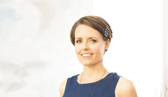 Professional woman with short brown hair and blue earrings smiling against a bright background.