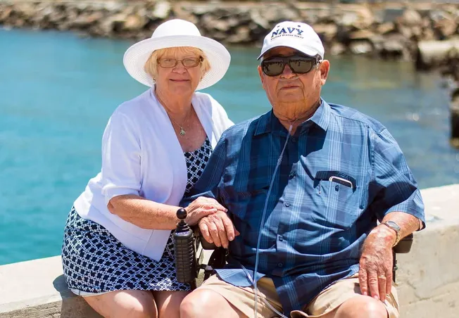 Enjoying retirement by the waterfront, senior couple with Navy veteran's hat, smiling and holding hands, sunny day, relaxing