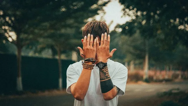 Colorful tattooed man covering his face with hands, standing outdoors in a park or street setting.