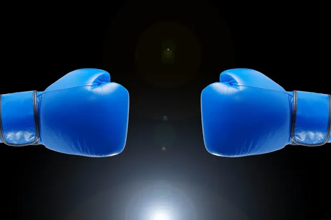 Blue boxing gloves facing each other against a black background, symbolizing competition or conflict, digital image related t