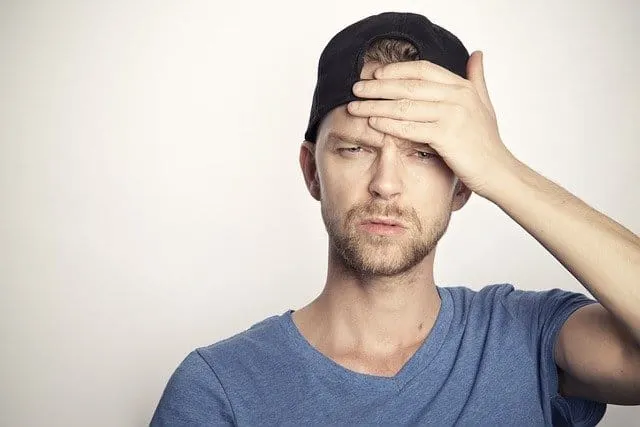 Frustrated young man holding his forehead, experiencing headache, stress or confusion, wearing a black cap and casual blue t-