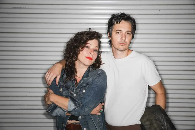 Woman and man standing close together in front of a metal garage door, casual style, music duo, indie artists, urban backgrou