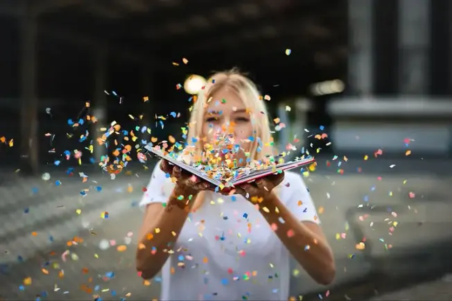 Bright young woman celebrating with colorful confetti and an open book outdoors, vibrant and joyful, illustrating excitement,