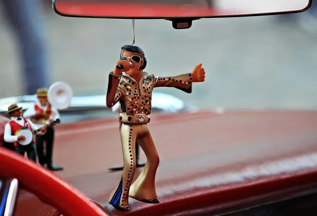 Retro Elvis Presley air freshener hanging from car rearview mirror with mini band figurines in the background.