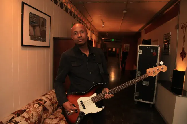 A musician with a bass guitar standing in a warmly lit, cozy venue, ready to perform or practice, surrounded by framed concer