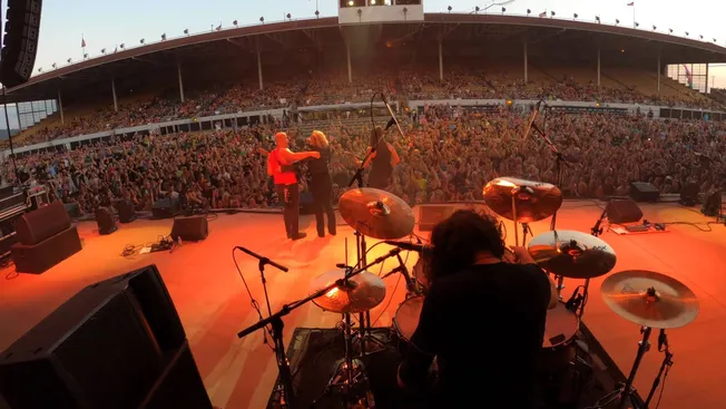 Vibrant live music performance at an outdoor concert with a large, enthusiastic audience enjoying the show. The stage feature