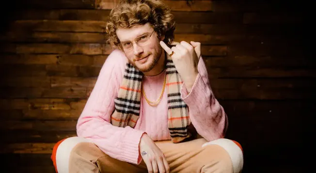 Young man with curly hair and glasses, wearing a pink sweater and a Burberry plaid scarf, sitting in front of a wooden backgr