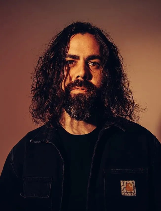 Portrait of a man with long curly hair and a beard, wearing a black denim jacket, against a warm-toned background, highlighti