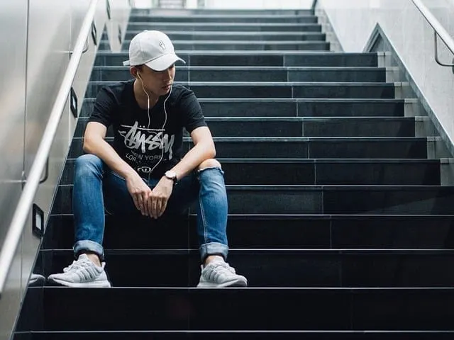 Youthful male sitting on city staircase, listening to music with earphones, casual urban style.