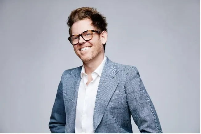 Fashionable man in gray blazer with glasses smiling in studio portrait - Hypebot.