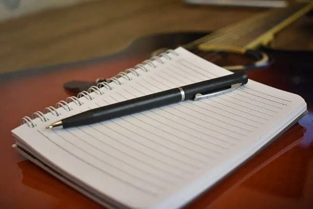 A notepad with lined pages and a pen resting on top, placed on a wooden desk with a guitar in the background.