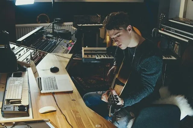 Music studio with young musician playing acoustic guitar surrounded by mixing consoles and electronic music equipment, creati