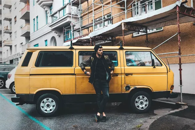 Yellow vintage van parked on city street with man leaning against it, in urban scene with apartment buildings and scaffolding