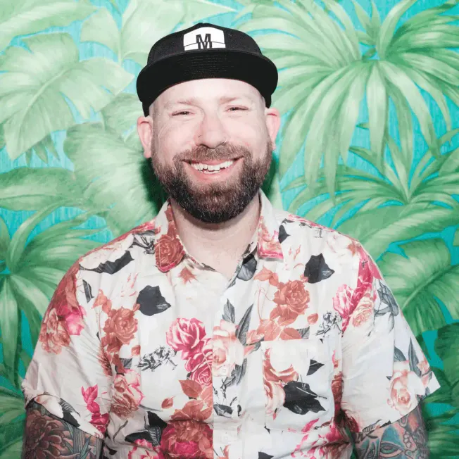 Smiling man wearing a floral shirt and cap in front of tropical leaf background, vibrant and lively ambiance, cheerful expres