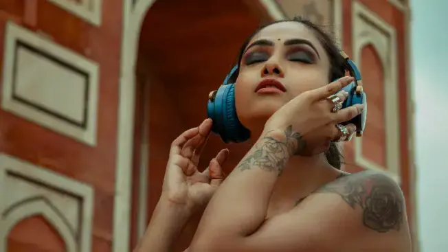 Headshot of a woman with tattoos enjoying music with blue headphones, standing outdoors against a historic brick building, ca